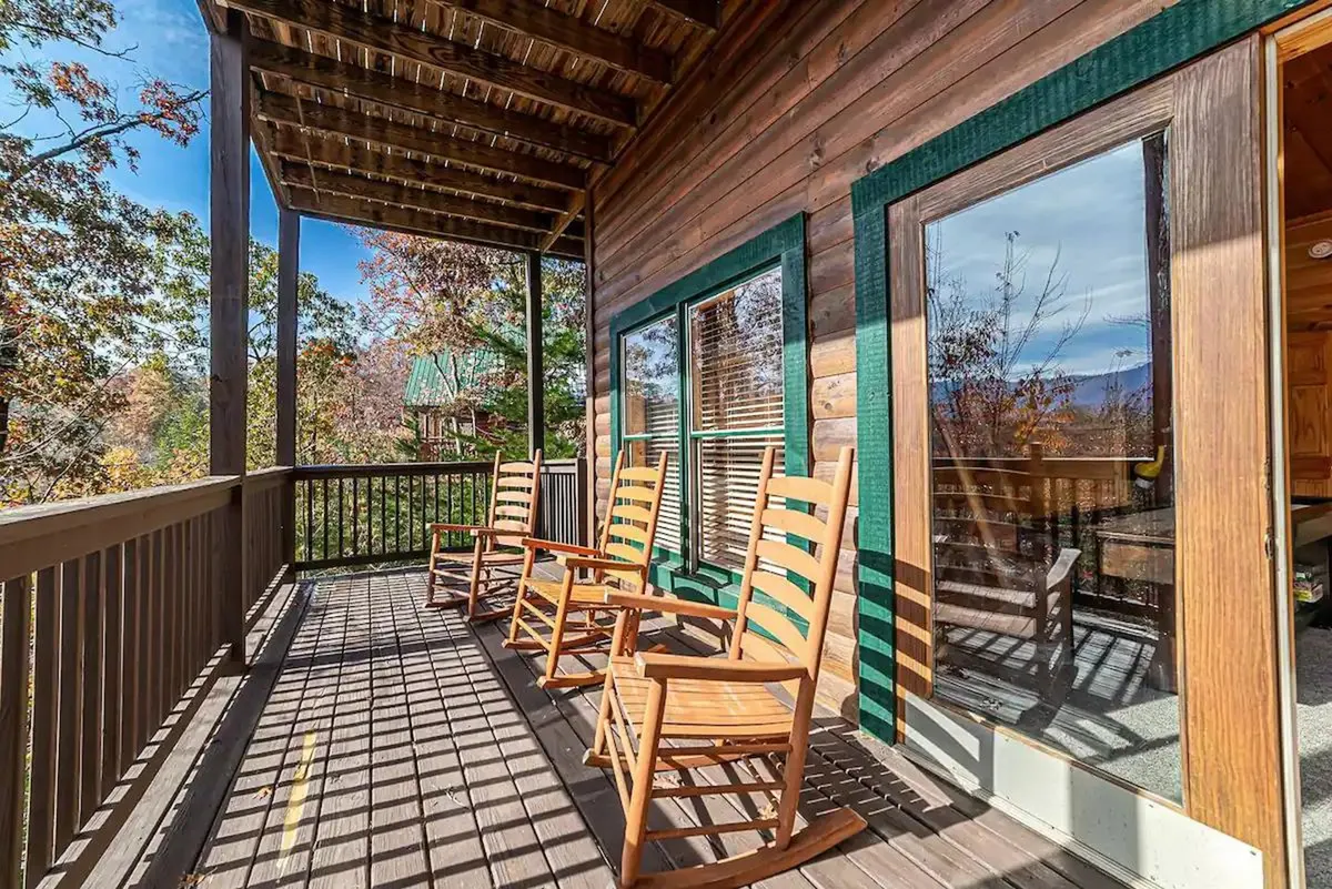 A spacious wooden deck lined with classic rocking chairs overlooks a serene natural setting. Large windows connect the deck to the cabin’s interior, offering a seamless transition between social indoor spaces and the outdoors. The deck is inviting and perfectly suited for group gatherings, relaxation, or stargazing after a day of adventures.