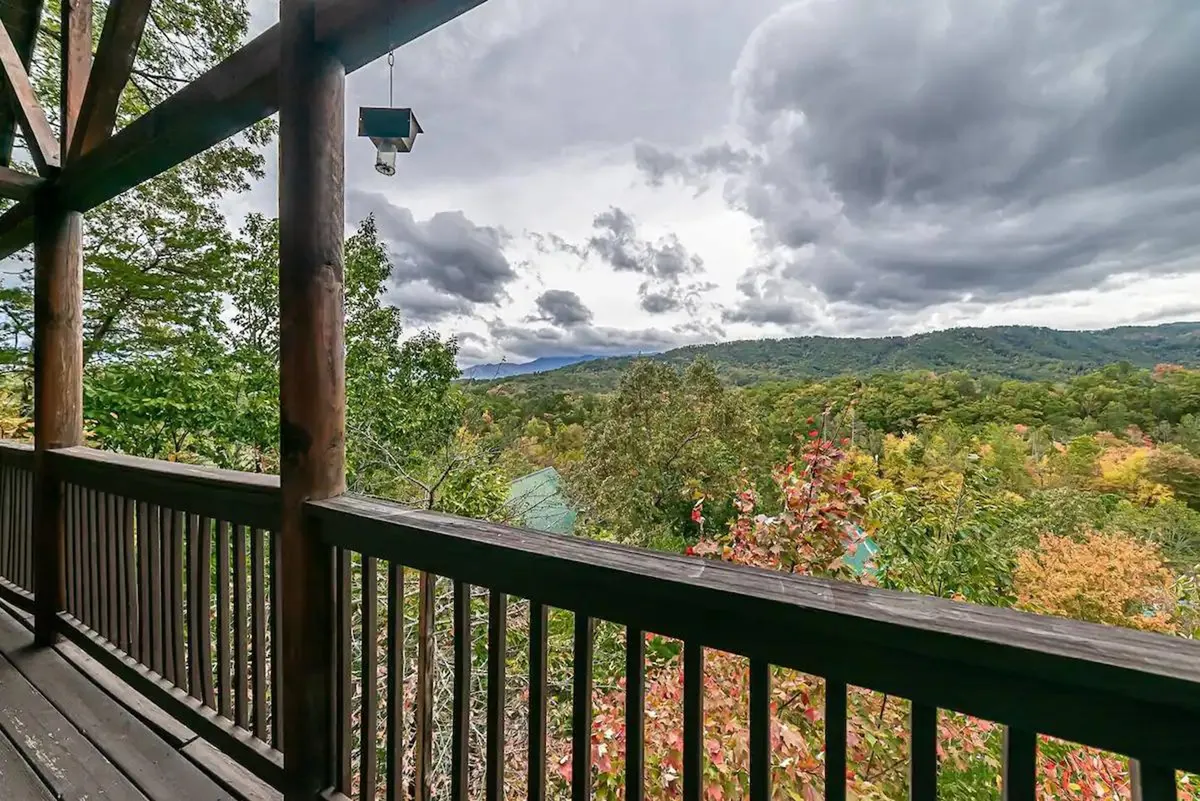 A view from a wooden deck, featuring a sturdy railing and offering an expansive vista of lush greenery and distant mountains. The scene conveys a calm, peaceful mood and emphasizes the beautiful natural views accessible from the deck.
