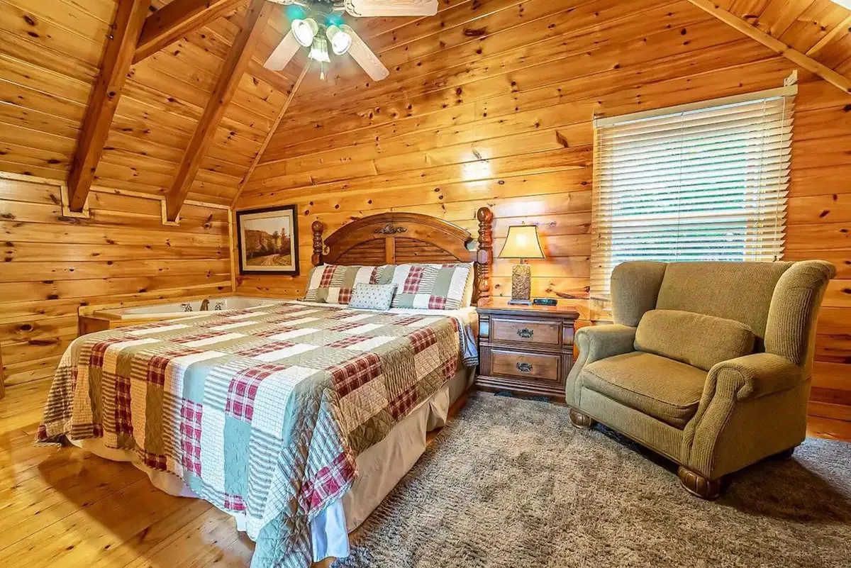 A cozy bedroom with a wooden ceiling, a plaid quilt on the bed, an armchair for reading, and a nightstand with a warm lamp beside the bed. The window allows for natural light and a glimpse of the outdoors, adding to the room's peaceful ambiance.