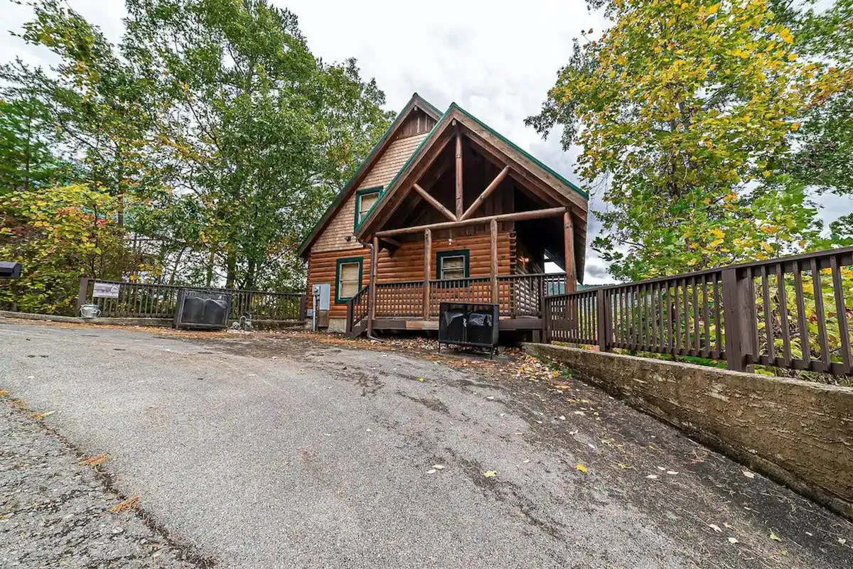 A classic log cabin nestled among mature trees, featuring a spacious front porch with seating for relaxing and gathering. The natural setting highlights the tranquility and natural beauty of the area, inviting families to unwind outdoors after a day of exploring Pigeon Forge's attractions.
