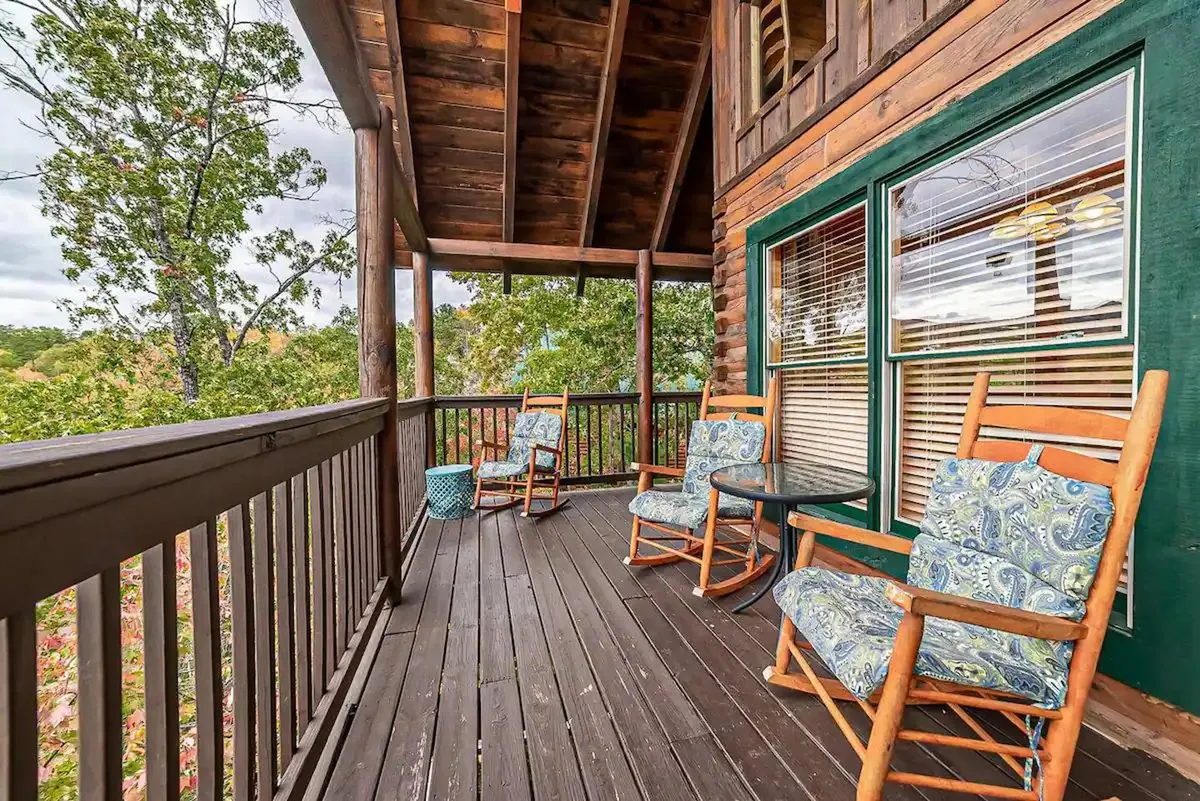 A cozy cabin deck furnished with classic wooden rocking chairs and a small table, looking out over natural surroundings. The deck's wooden railings blend seamlessly with the tranquil outdoor setting, inviting guests to relax, read, or simply enjoy the gentle sounds of nature.