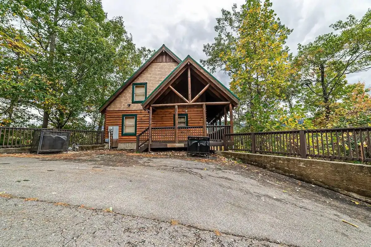 The image features a charming log cabin nestled among lush trees, with a spacious front porch perfect for relaxing and enjoying the mountain surroundings. The setting exudes warmth and tranquility, showcasing the cabin’s inviting exterior and the welcoming natural beauty of Pigeon Forge.