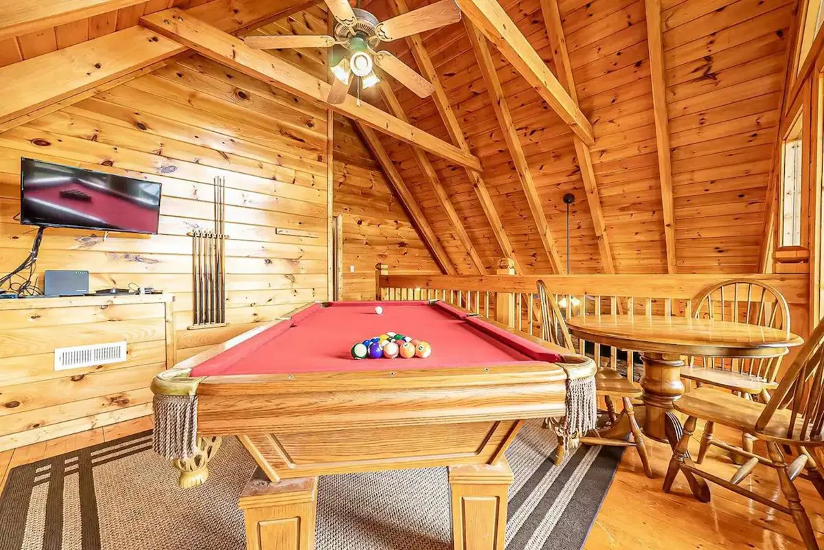 A vibrant living space with a pool table at the center, wooden ceiling beams overhead, a dining table nearby, and a television on the wall. People are engaged in playing pool, watching TV, and socializing, creating a lively and fun environment.