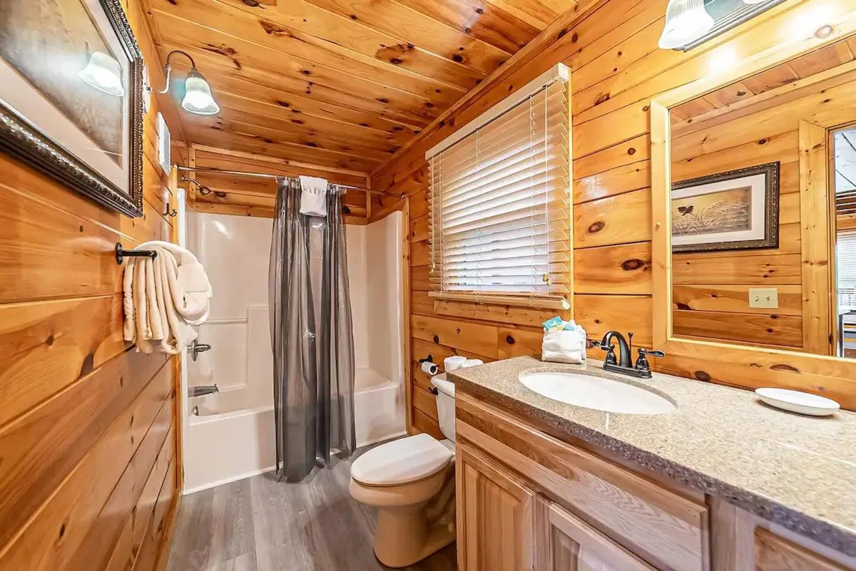 A rustic bathroom with wooden paneling, a granite countertop, a large mirror, and a deep soaking tub. The ambiance is calming and inviting, ideal for relaxation and self-care routines.