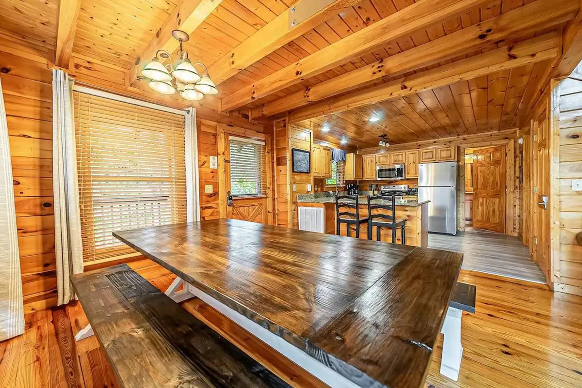 A spacious interior combining a large wooden dining table with sturdy wooden beams and walls, seamlessly connecting the dining area to the adjacent kitchen with visible appliances. The arrangement sets the scene for family meals, celebrations, and group gatherings.