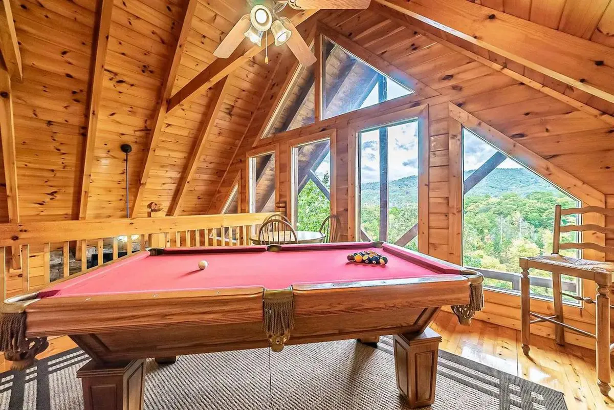 A cozy indoor game area features a pool table, wooden furniture, and large windows with mountain views. The space is designed for group entertainment, with ample seating for friends to gather, socialize, and enjoy playful competition.